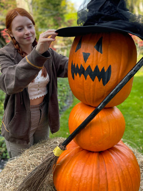 close-up image of attractive, red-haired woman arranging a witch's hat on pumpkin witch, three orange gourds stacked to make 'witch' with broomstick, surrounded by hay, halloween face jack o'lantern outdoors on lawn, focus on