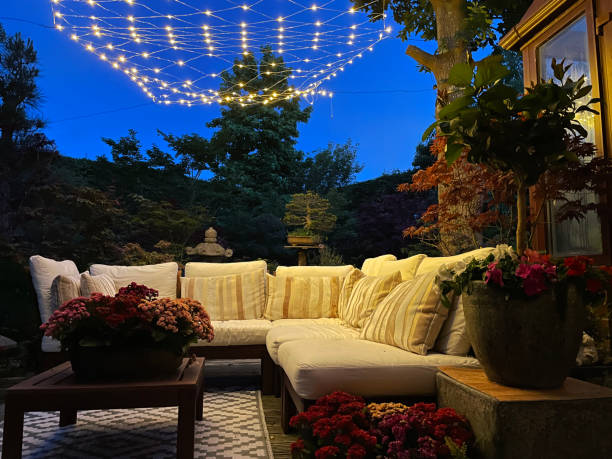 close-up image of outdoor lounging area at night illuminated by string fairy lights, hardwood seating with cushions, wooden table top with flowering plant centrepiece, bonsai trees, japanese maples, landscaped oriental design
