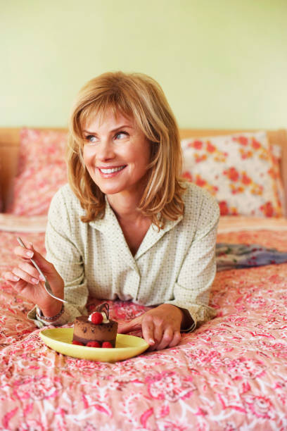 close-up of a mature woman lying on the bed and eating cake - junk food stockfoto's en -beelden