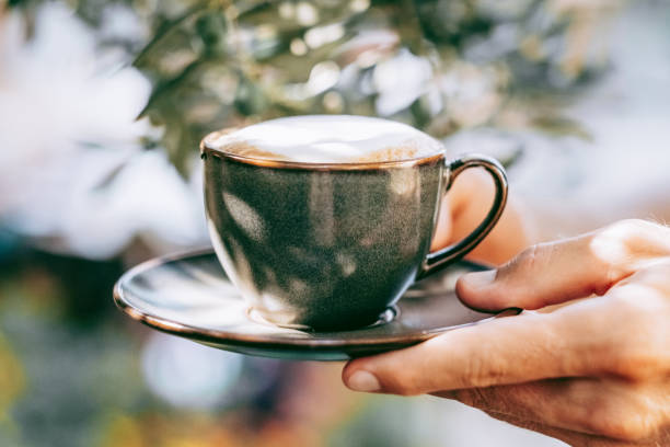 close-up of hands holding a cup of cappuccino in a sunlit garden, with soft focus greenery in the background. concept of enjoying coffee outdoors. - garden decoration stock pictures, royalty-free photos & images