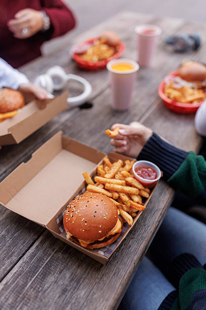 close up of hand holding french fries, there is a box with burger on a table - junk food stock pictures, royalty-free photos & images