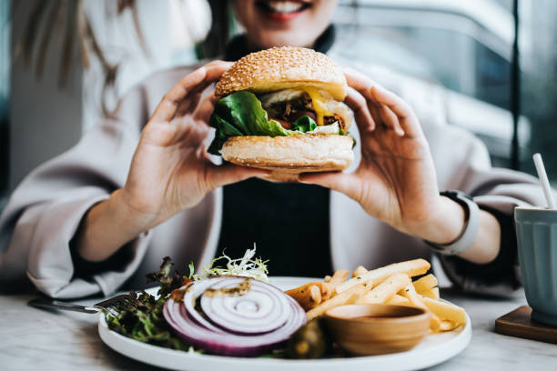 close up of happy young asian woman sitting by the window in a restaurant, enjoying lunch during the day. she is having freshly made delicious burger with fries and side salad. lifestyle, people and food concept - junk food s