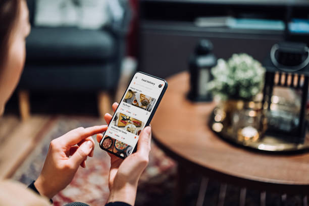 close up of young asian woman using meal delivery service and ordering food online with mobile app on smartphone while sitting on the sofa in the living room at a cozy home - junk food stock pictures, royalty-free photos & im