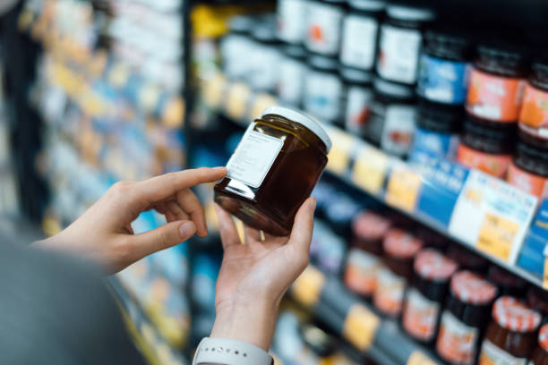 close up of young woman grocery shopping in a supermarket. standing by the aisle, holding a bottle of marmalade, reading the nutritional label and checking ingredients at the back - food fotografías e imágenes de stock