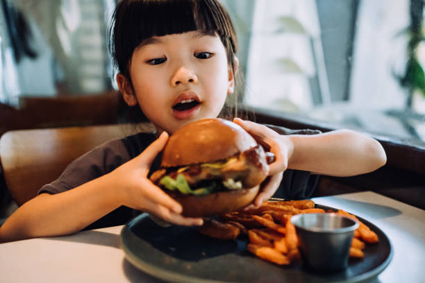 close up shot of funny little asian girl looking excited while holding a big burger and making a face while enjoying eating burger with sweet potato fries in a restaurant. eating out lifestyle - junk food stock pictures, roya