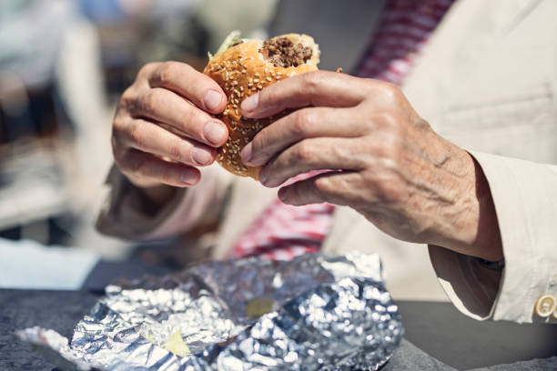 closeup of senior man eating a burger outdoors on a sunny day. - junk food stock pictures, royalty-free photos & images