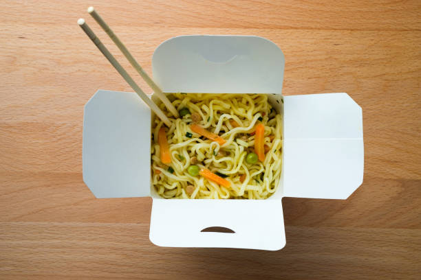 cooked instant noodles, sprinkled with spices, vegetables and herbs, freeze-dried meat. nearby are wooden chopsticks. asian chicken soup with ramen noodles in a paper box, on the dining room kitchen table. the concept of home