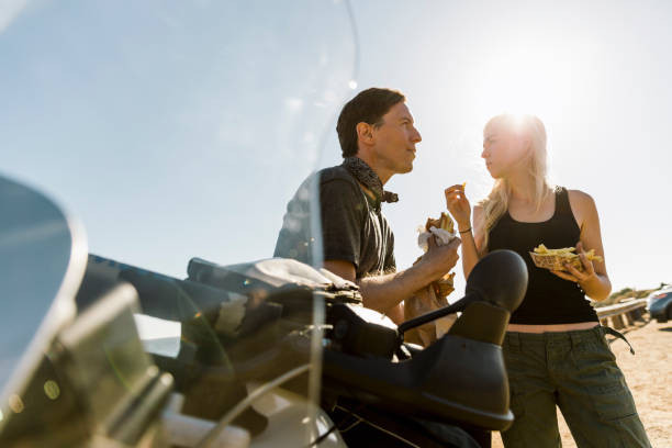 couple enjoying meal by motorcycle against sky on sunny day - junk food stock pictures, royalty-free photos & images
