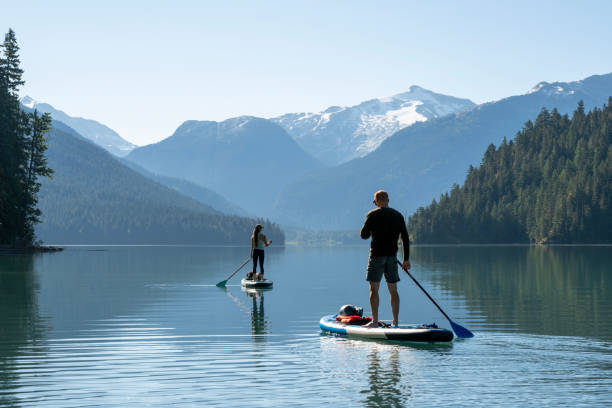 couple paddle sup boards across mountain lake, whistler - travel stock pictures, royalty-free photos & images