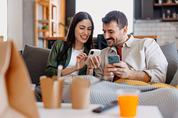 couple relaxing on sofa using smartphones and eating takeaway food - junk food stock pictures, royalty-free photos & images