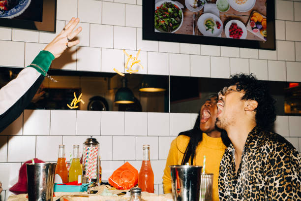 cropped hand of man throwing french fries at male and female friends with open mouth in cafe - food stock pictures, royalty-free photos & images
