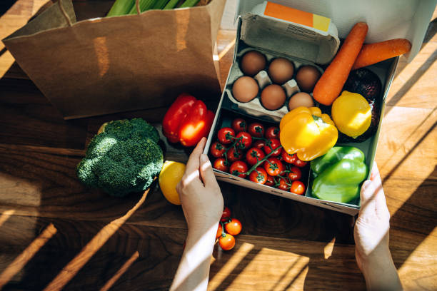 cropped hands of woman opening a home delivery cardboard box filled with multi-coloured fresh organic fruits, vegetables and groceries on wooden background against sunlight. grocery subscription boxes. meal kits. convenient a