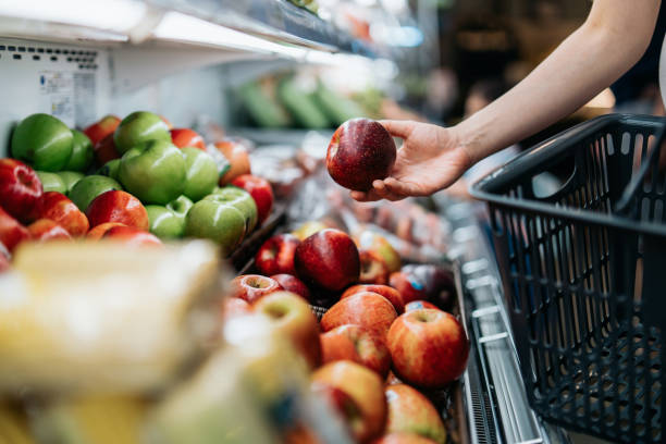 cropped shot of young asian woman choosing fresh organic fruits in supermarket. she is picking a red apple along the produce aisle. routine grocery shopping. healthy living and eating lifestyle - food stock pictures, royalty-
