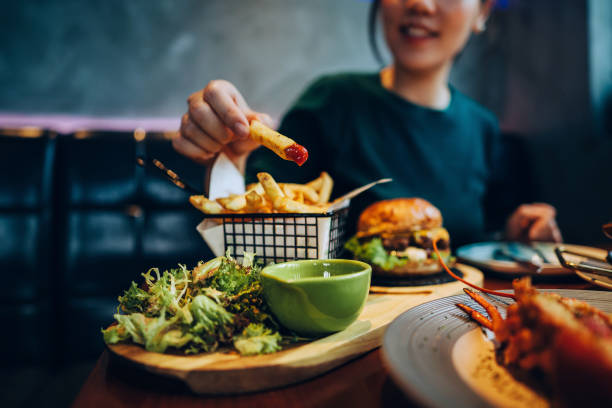 cropped shot of young asian woman eating freshly made delicious burger, dipping fries in ketchup on the dining table. enjoying a feast in the restaurant. people, food and lifestyle - junk food stock pictures, royalty-free pho