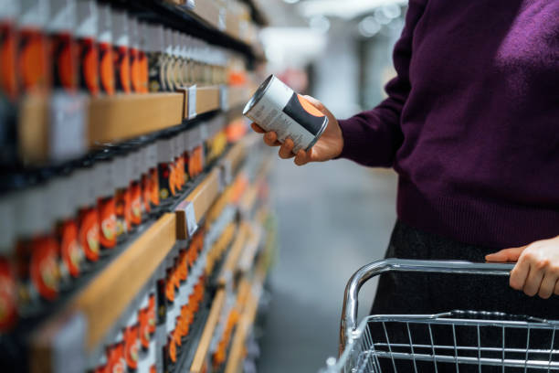 cropped shot of young woman pushing a shopping cart, grocery shopping in supermarket. she is holding a tin can and reading the nutritional label. routine grocery shopping. food shopping. making healthier food choices - junk f