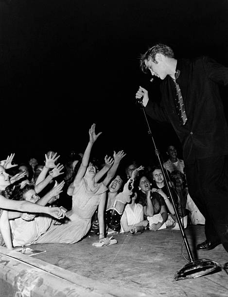 Crowd of screaming teenage girls reach for Elvis Presley as he sings on stage.