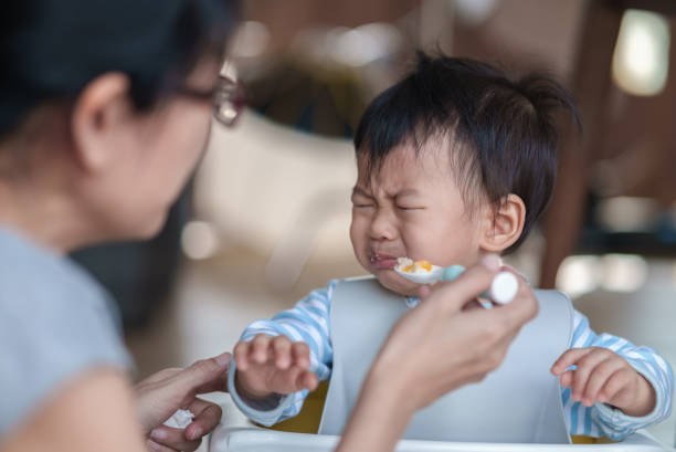 cute baby boy refusing to eat food and crying while his mother feeds him. - food stock pictures, royalty-free photos & images