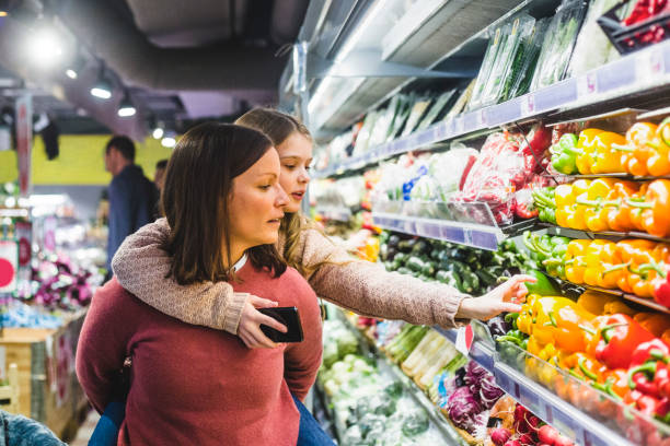 cute daughter choosing bell peppers while being piggybacked by mother in store - food fotografías e imágenes de stock