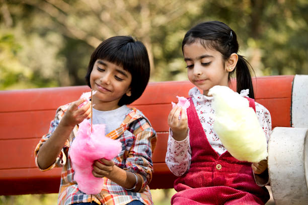 cute siblings eating cotton candies while sitting in park - junk food stock pictures, royalty-free photos & images