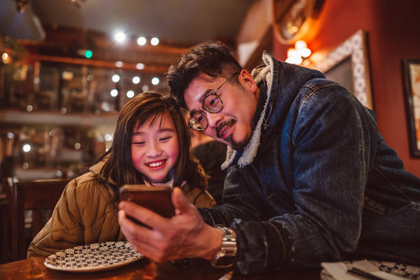 dad & daughter using smartphone in restaurant joyfully - food fotografías e imágenes de stock