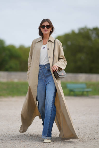 Darja Barannik wears sunglasses, a white top, a beige long trench coat, blue denim jeans / pants, outside Loewe, during the Womenswear Spring/Summer...