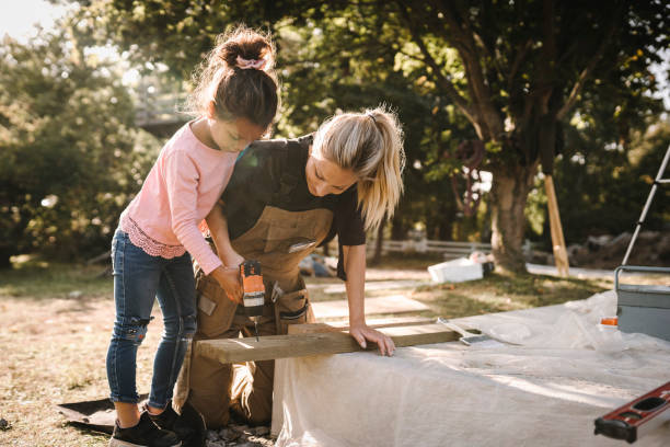 daughter using drill machine on wooden plank by mother in backyard - home decoration stockfoto's en -beelden