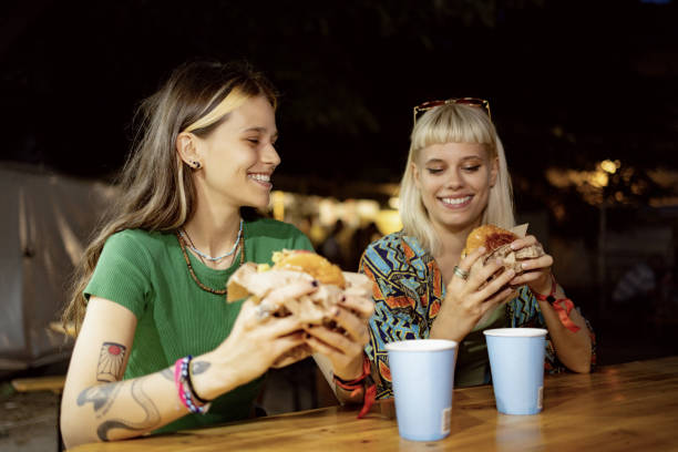 des femmes heureuses mangeant des hamburgers lors d’un festival de musique la nuit. - concert photos et images de collection