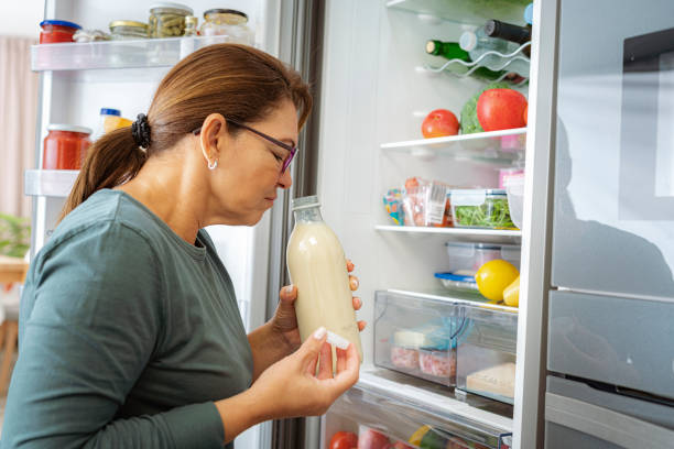 disgusted woman smelling expired milk by the fridge - food stock pictures, royalty-free photos & images
