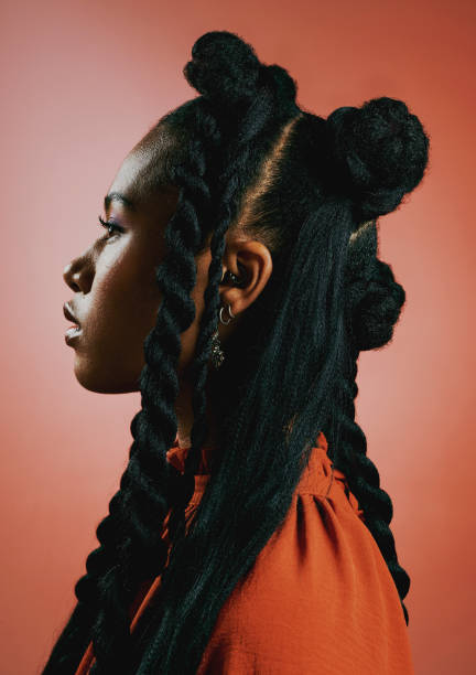edgy young black woman with trendy braided hairstyle against an orange studio background. profile of a confident, serious and stylish african american female with bold personality and cool attitude - hairstyle stockfoto's en 