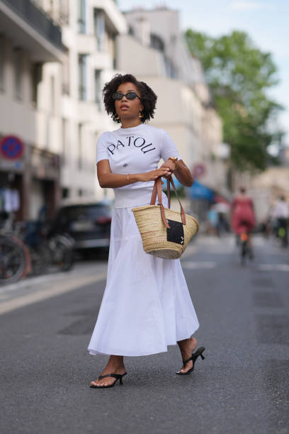 Ellie Delphine wears black sunglasses, gold earrings, white short sleeve Patou shirt, gold bracelets, white high waisted flowy maxi skirt, dark brown...