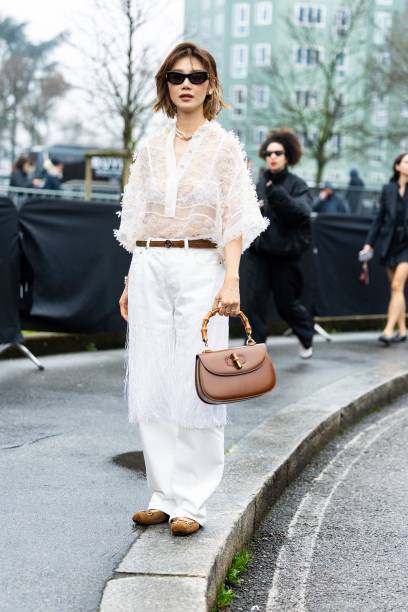 Elva Ni wears black sunglasses, a gold necklace, a white see-through long sleeved flower shirt, a white bra, a brown leather Gucci belt, a brown...