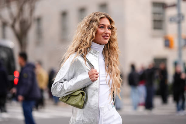 Emili Sindlev wears earrings, a silver shiny jacket, a white turtleneck top, a green leather Coach bag, white pants with printed green apples,...