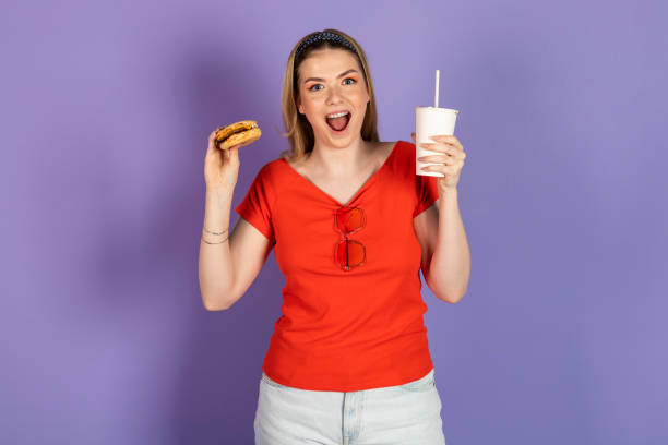 excited hungry pretty girl eats fast food, cheap burger, holds a cheeseburger and soda, open mouth. fast food and unhealthy diet. isolated on purple background. - junk food stock pictures, royalty-free photos & images