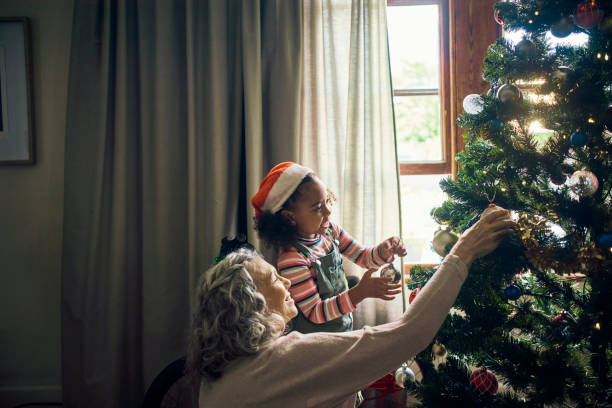 family, christmas tree and decor with a mature woman and child preparing for festive season with love. grandmother, granddaughter and decorating an xmas tree with ornaments and bauble in family home - home decoration stock pi