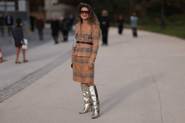 Fashion week guest is seen wearing an orange and green plaid bouclé jacket with a black Chanel belt, a knee-length skirt, silver high boots and brown...