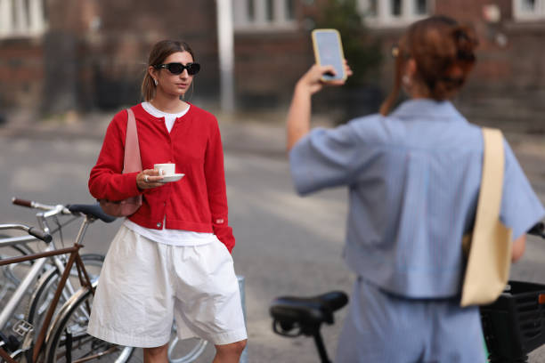 Fashion Week guest is seen wearing black sunglasses from Ganni, several silver hoop earrings, a red buttoned cardigan, a white t-shirt underneath, a...