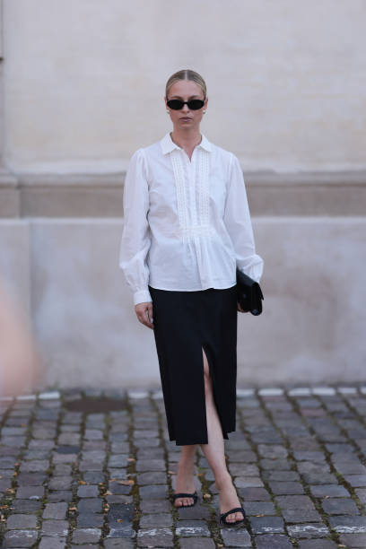 Fashion Week guest is seen wearing oval black sunglasses, silver round earrings, a long-sleeved white blouse with lace details at the chest, collar...