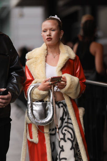 Fashion Week Guest was seen wearing white pants with black writings, a white cropped shirt, a silver bag that is shiny along with lots of jewelry and...