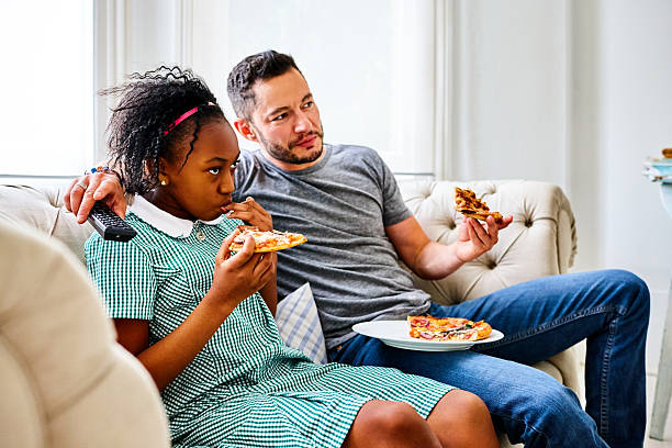 father and daughter eating pizza and watching tv - junk food stockfoto's en -beelden