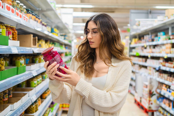 female professional studying product label, wearing eyeglasses in grocery store aisle, surrounded by packaged foods, comparing nutritional details - food stock pictures, royalty-free photos & images