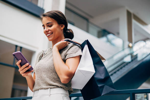 femme heureuse faisant du shopping et utilisant un smartphone dans un centre commercial moderne - fashion photos et images de collection