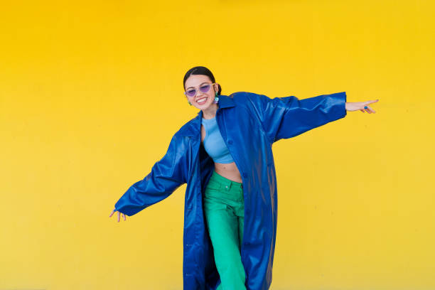 femme élégante posant en manteau bleu, lunettes de soleil foncées sur fond jaune vif, rayonnant de confiance urbaine contemporaine - fashion photos et images de collection