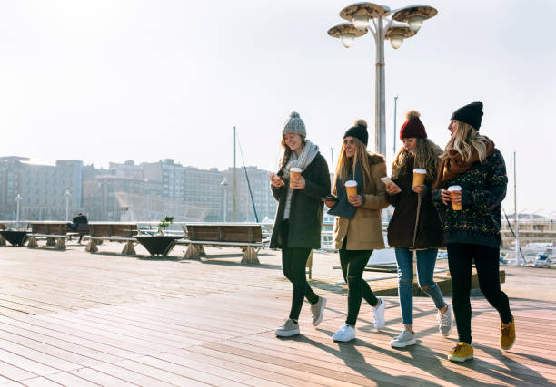 four friends with coffee to go and cell phones walking on promenade in winter - junk food stock pictures, royalty-free photos & images