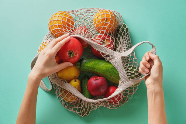 fresh juicy fruits and vegetables, products in a reusable shopping bag. a girl or woman takes or lays out products from a string bag made from recycled materials on a green pastel background. vegetarianism, veganism. no plast