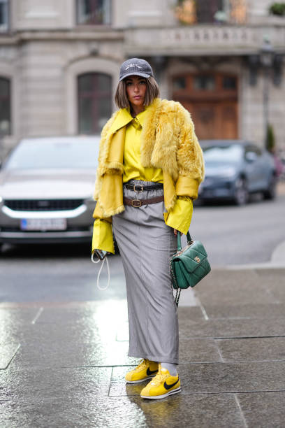 Gabriella Berdugo wears a cap hat, a yellow oversized shirt, a fluffy winter faux fur jacket, two leather belts, a gray long skirt, a green Chanel...