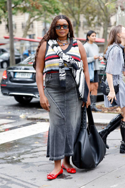 Gabriella Karefa Johnson wears striped knitted vest, grey long denim skirt, black tote bag, red ballet flats, outside Ganni, during Womenswear...