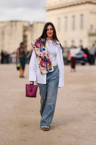 Gala Gonzalez wears white button up long sleeve shirt, navy blue denim jean pants, dark brown leather boots, dark red leather bag, outside Loewe,...