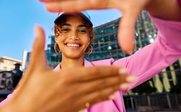 gen z woman framing her face with her hands and smiling. close-up shot with modern building in the background. positive emotions and urban lifestyle concept. - fashion stock pictures, royalty-free photos & images