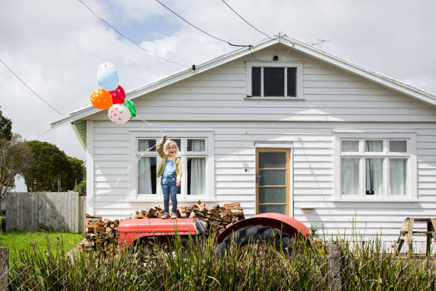 girl stands on a tractor holding colourful balloons in front of a bungalow - garden decoration stock pictures, royalty-free photos & images