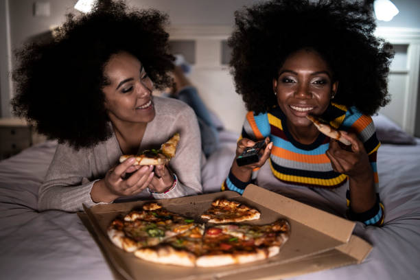 girlfriends lying down on bed and eating pizza - junk food stock pictures, royalty-free photos & images